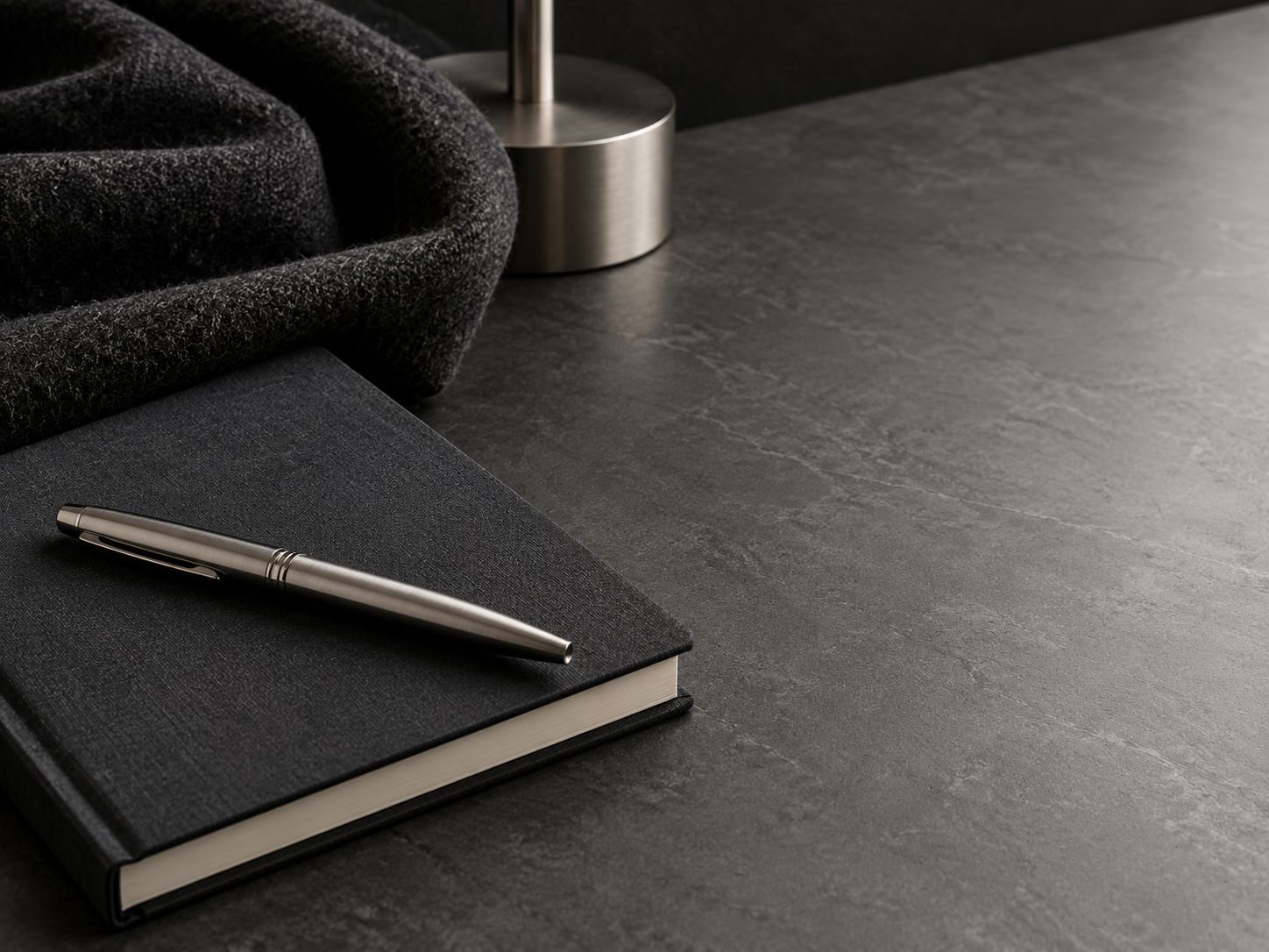 A closed black notebook and a pen on a dark desk, with a folded wool cloth and a metal lamp.